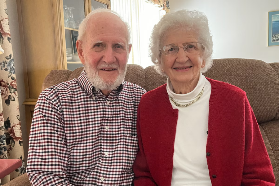 An elderly couple sits closely together on a beige recliner in a cozy living room. The man wears a checkered shirt and has a white beard, while the woman wears glasses, a red cardigan over a white turtleneck, and a gold chain necklace, both smiling warmly at the camera.