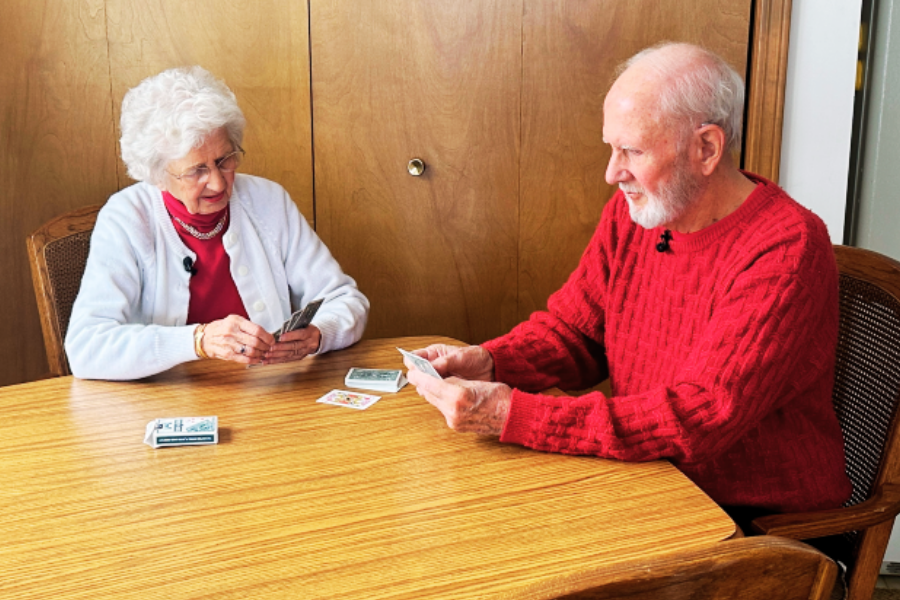 An elderly couple sits across from each other at a wooden table, playing a card game. The woman wears a white cardigan over a red top and pearl necklace, while the man wears a red knit sweater, both smiling and focused on their cards.