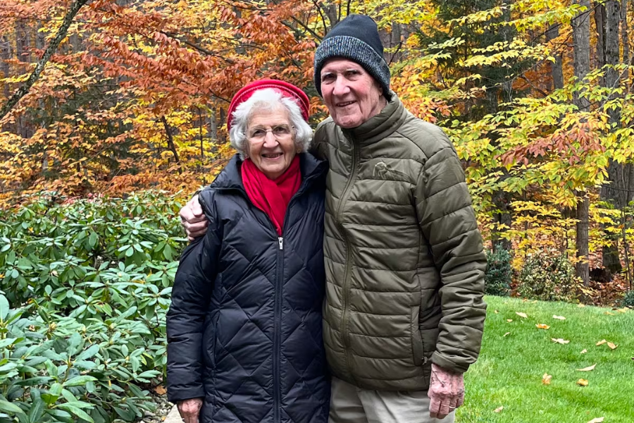 An elderly couple bundled in winter jackets poses happily in front of a backdrop of vibrant fall foliage. The woman wears a black coat, red scarf, and red hat, while the man sports a green puffer jacket and a knit beanie, standing on a green lawn surrounded by colorful autumn trees.