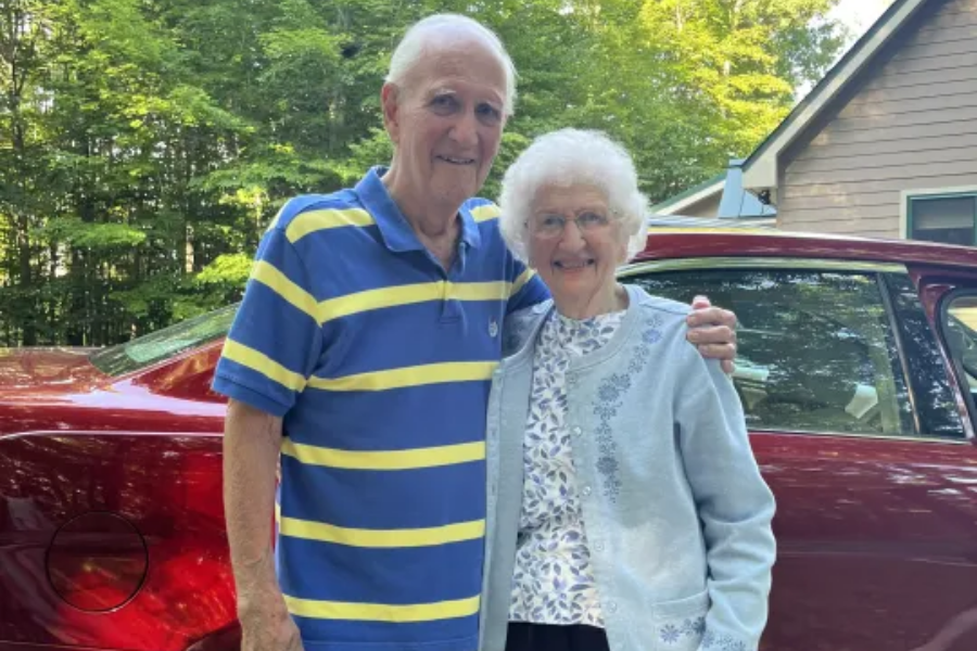 An elderly couple stands smiling with arms around each other in front of a shiny red car. The man wears a blue and yellow striped polo shirt, and the woman wears a light blue cardigan with floral embroidery; a wooded area and house are visible in the background.