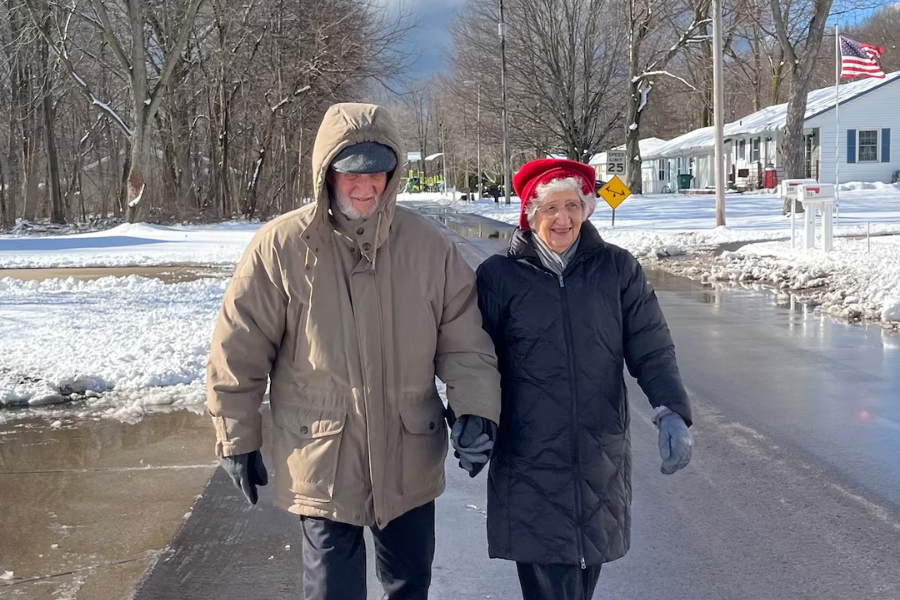 An elderly couple bundled in winter coats walks hand-in-hand down a snowy street on a sunny day. The man wears a tan parka with the hood up and black gloves, while the woman wears a black coat, red hat, and gray gloves, smiling as they enjoy the winter stroll.