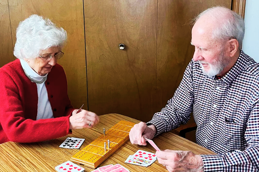 An elderly couple sits at a wooden table playing cribbage, focused on the game. The woman, dressed in a red cardigan and white turtleneck, is moving a peg on the cribbage board, while the man in a plaid shirt holds playing cards and smiles.