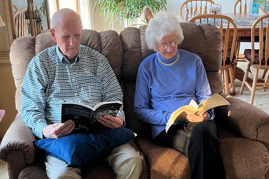 An elderly couple sits side by side on a brown recliner, each reading a book in a cozy living room. The man wears a green checkered shirt and rests a paperback on a blue pillow, while the woman wears a periwinkle top and pearl necklace, smiling gently as she reads.