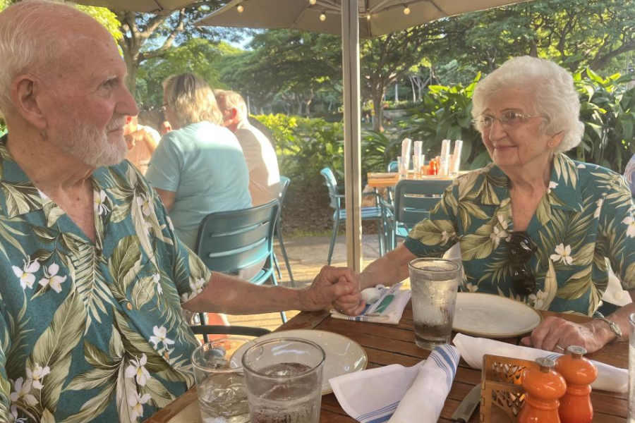 An elderly couple sits at an outdoor restaurant table, holding hands and smiling warmly at each other. Both wear matching tropical floral shirts, and their table is set with plates, glasses of water, and silverware under a sunlit patio umbrella with greenery in the background.