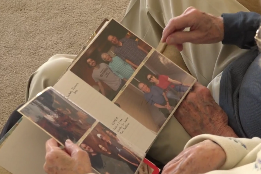 Close-up of an elderly person's hands holding a photo album open on their lap, displaying several family photos and handwritten captions. The album rests on beige pants, and another person with light skin and a floral sweater is seated beside them, gently turning the page.