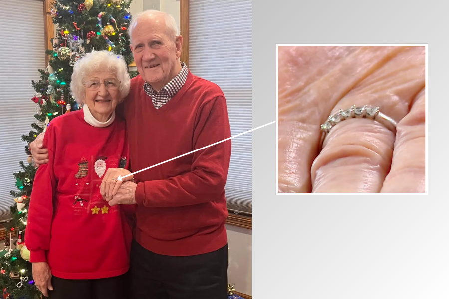 An elderly couple stands smiling in front of a decorated Christmas tree, both dressed in festive red sweaters. The man gently holds the woman’s hand, which features a close-up inset of a diamond anniversary band with five stones, highlighting a special moment of love and commitment.