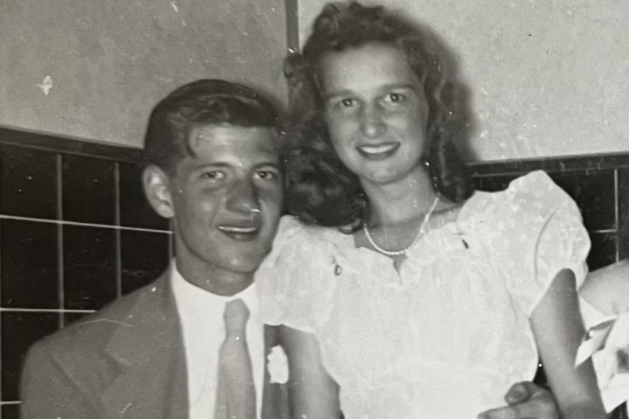 A black-and-white vintage photo of a young couple smiling together, dressed in formal attire. The man wears a suit and tie with a boutonnière, while the woman wears a puff-sleeved white dress and a delicate necklace, with wavy dark hair cascading over her shoulders.