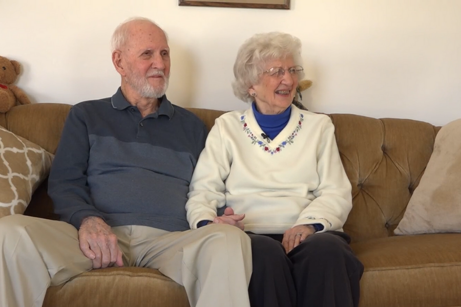 An elderly couple sits close together on a tan sofa, holding hands and smiling. The man wears a dark polo shirt and beige pants, while the woman wears glasses, a white sweater with floral embroidery, and a blue turtleneck underneath; cozy pillows and a teddy bear are visible in the background.