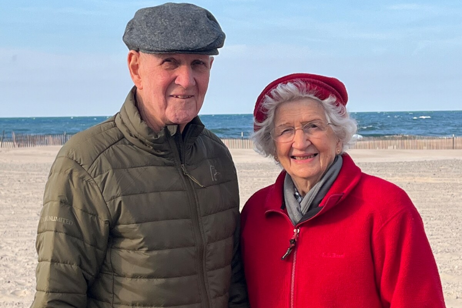 An elderly couple poses warmly on a sandy beach with the ocean in the background. The man wears a green quilted jacket and a gray flat cap, while the woman is bundled in a bright red jacket and matching beret, smiling with the waves behind them.