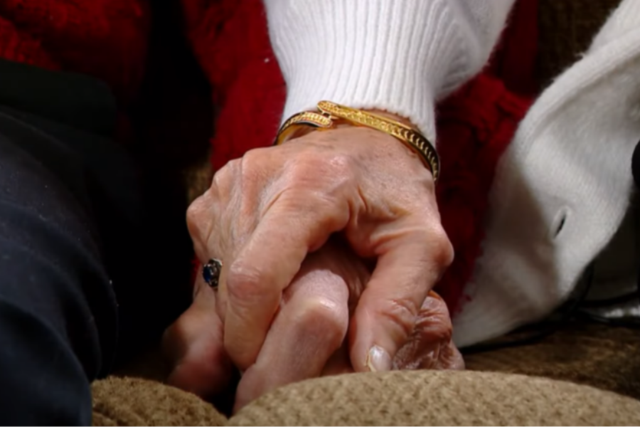 Close-up of two elderly hands gently clasped together, resting on a couch. One hand wears a gold bracelet and a dark gemstone ring, conveying a tender moment of love and connection.