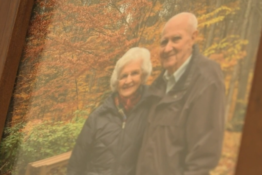 A framed photo of an elderly couple standing close together and smiling, set against a vibrant backdrop of autumn trees with orange and yellow leaves. Both wear jackets, suggesting a crisp fall day.