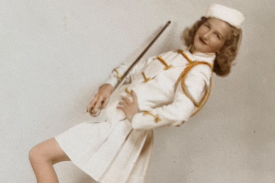 A young woman poses confidently in a vintage white and gold majorette uniform, holding a baton and smiling toward the camera. She wears a pleated skirt, fitted jacket with gold trim, and a matching white cap, striking a playful marching stance.