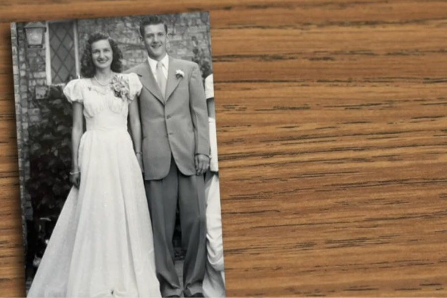 A black-and-white vintage photograph of a young couple dressed for a formal occasion, possibly prom or a wedding, is placed on a wooden surface. The woman wears a floor-length gown with puffed sleeves and a corsage, while the man wears a suit and boutonnière, both smiling brightly.