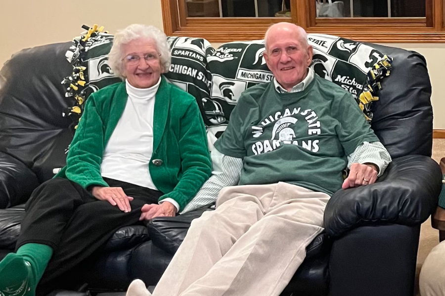 An elderly couple sits on a black leather couch, smiling and dressed in Michigan State Spartans gear. The woman wears a green jacket over a white turtleneck and green socks, while the man sports a green Spartans T-shirt over a striped shirt. A Michigan State fleece blanket is draped over the couch behind them.