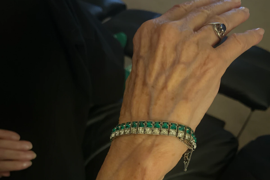 Close-up of an elderly woman's hand wearing a silver bracelet with alternating square-cut emerald and clear stones. She also wears a silver ring with a blue gemstone, and the background is softly out of focus, showing part of a black outfit.
