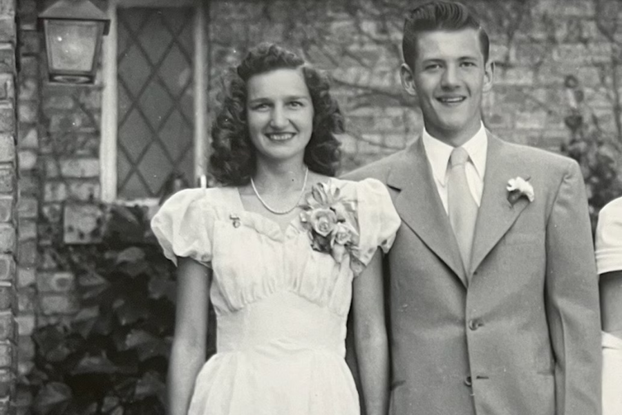 A black-and-white photo of a young couple dressed formally and smiling, standing in front of a brick house with leaded glass windows. The woman wears a light-colored dress with puffed sleeves and a large floral corsage, while the man wears a suit with a boutonnière and a light tie.