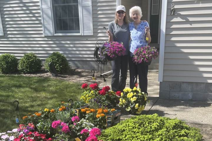 Two women stand smiling in front of a house, each holding a hanging basket filled with vibrant purple flowers. They are surrounded by a colorful assortment of potted flowers on the ground, including yellow, orange, pink, and red blooms, ready for planting in the garden.