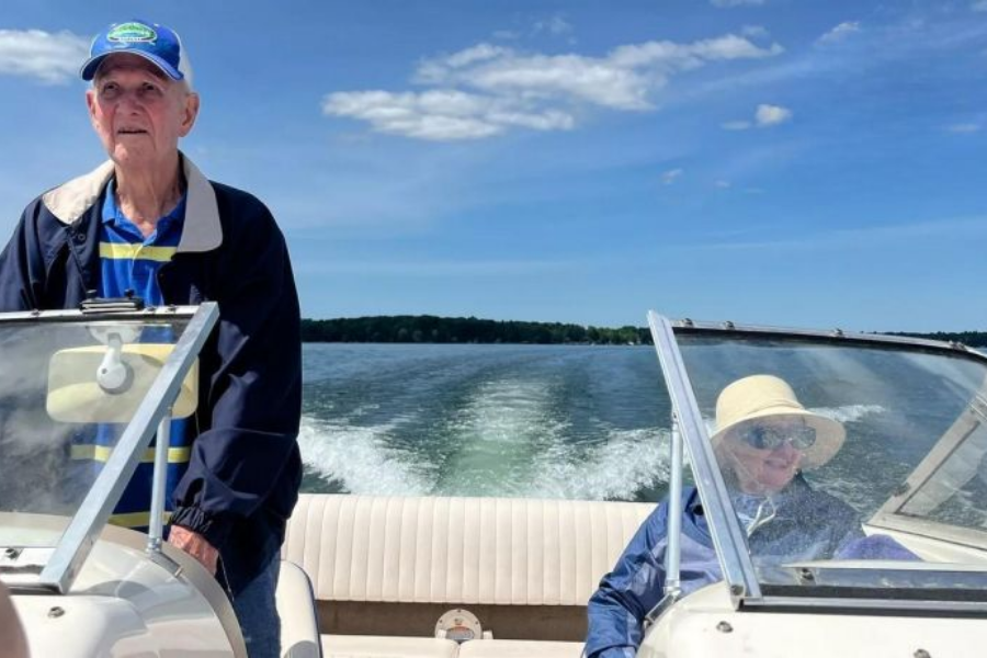 An elderly couple enjoys a sunny boat ride on a lake, with the man standing at the helm wearing a blue and yellow striped polo, a navy jacket, and a ball cap. The woman, seated and smiling, wears a wide-brimmed sunhat, sunglasses, and a light jacket, with a scenic wake trailing behind them on the water.