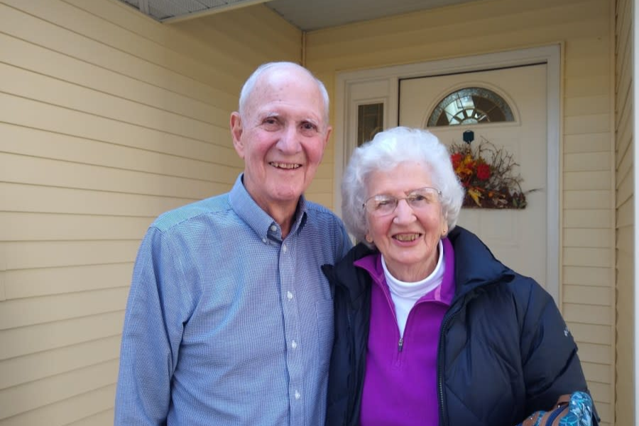 An elderly couple smiles warmly while standing on the porch of a house with light yellow siding. The man wears a blue checkered shirt, and the woman wears glasses, a purple top, and a black puffy jacket; a festive autumn wreath decorates the door behind them.
