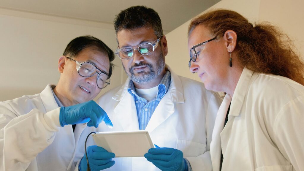 Three researchers in lab coats reviewing data on a digital tablet.