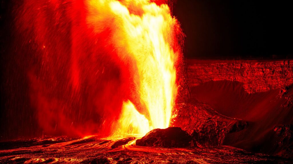 A dramatic volcanic eruption sends bright red and yellow lava spraying into the air against a dark background.