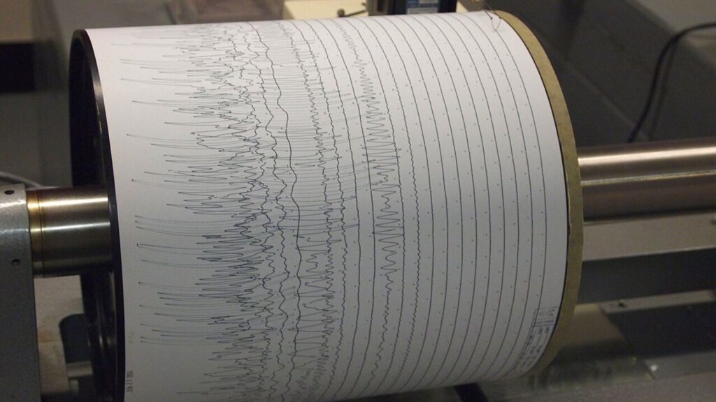 A seismograph drum records dense, jagged lines representing earthquake vibrations over time.