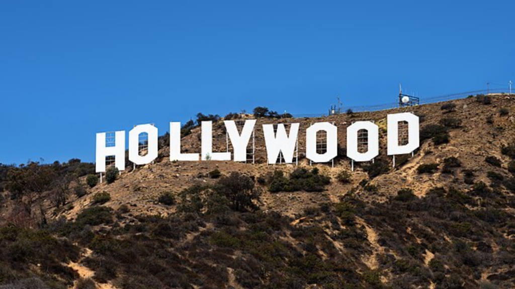 The famous white Hollywood sign stands on a dry, brush-covered hillside under a clear blue sky.