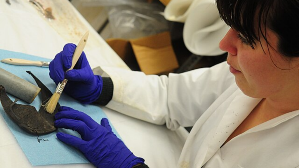 A person wearing blue gloves carefully cleans a dark artifact with a small brush.