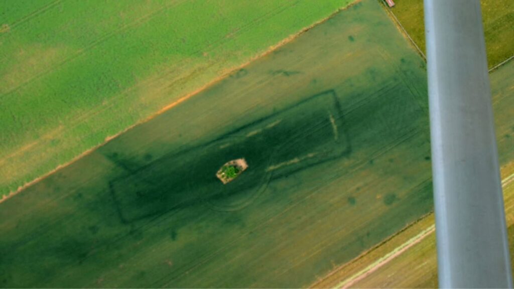 Aerial view of a green field with dark cropmarks forming a rectangular enclosure around a small central mound, likely a buried barrow.